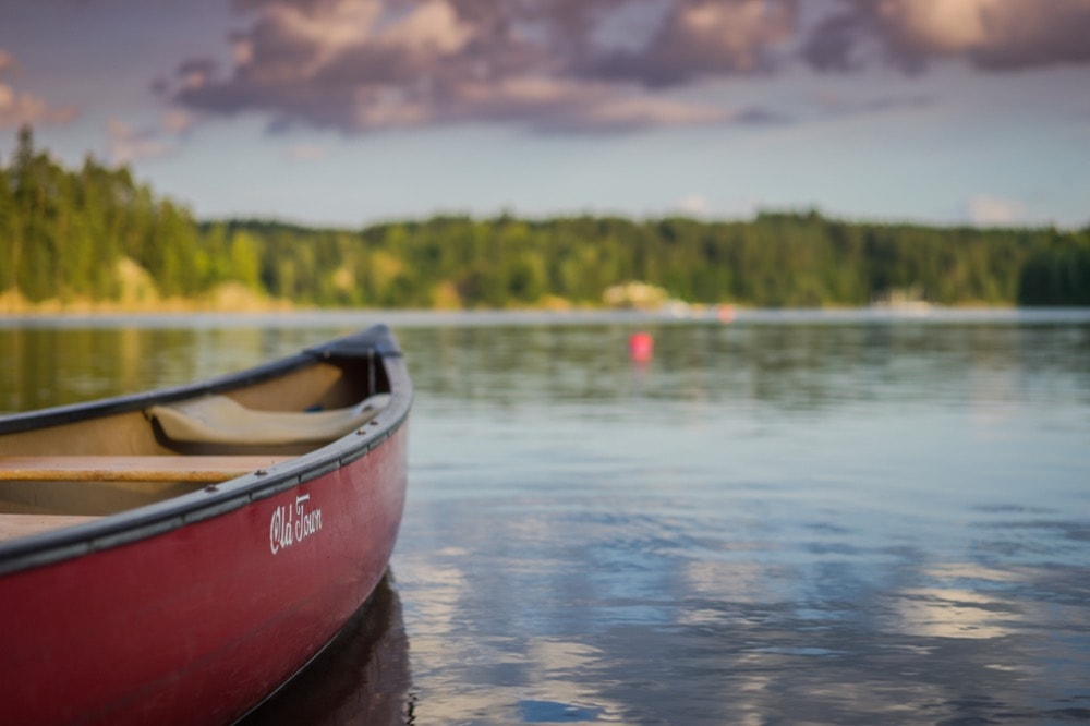 A close-up of the bow of a red canoe labeled "Old Town" gently resting on calm lake waters. In the blurred background, the serene landscape features a treeline and cloudy sky reflecting on the water's surface, with a buoy visible in the distance.
