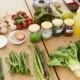 A wooden table filled with fresh ingredients, including leafy greens, asparagus, green beans, chives, mushrooms, cherry tomatoes, and herbs. Small bowls contain cheese, cream, and an egg. Jars of spices and seeds labeled "Sunflower," "Pumpkin," and "Paprika" are placed nearby. A yellow squeeze bottle, a green apple, and kitchen utensils are also present, suggesting a cooking or meal preparation setting.