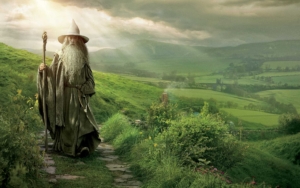 Gandalf the Grey from The Lord of the Rings stands on a stone path in the lush green hills of the Shire. Wearing his iconic pointed wizard hat and gray robes, he holds his wooden staff while overlooking the idyllic countryside. Sunlight breaks through clouds in the background, illuminating the pastoral landscape of rolling hills and distant fields that stretches toward the horizon. The scene captures Gandalf as the wise mentor figure at the beginning of the hero's journey.