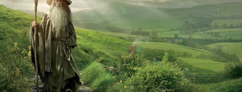 Gandalf the Grey from The Lord of the Rings stands on a stone path in the lush green hills of the Shire. Wearing his iconic pointed wizard hat and gray robes, he holds his wooden staff while overlooking the idyllic countryside. Sunlight breaks through clouds in the background, illuminating the pastoral landscape of rolling hills and distant fields that stretches toward the horizon. The scene captures Gandalf as the wise mentor figure at the beginning of the hero's journey.