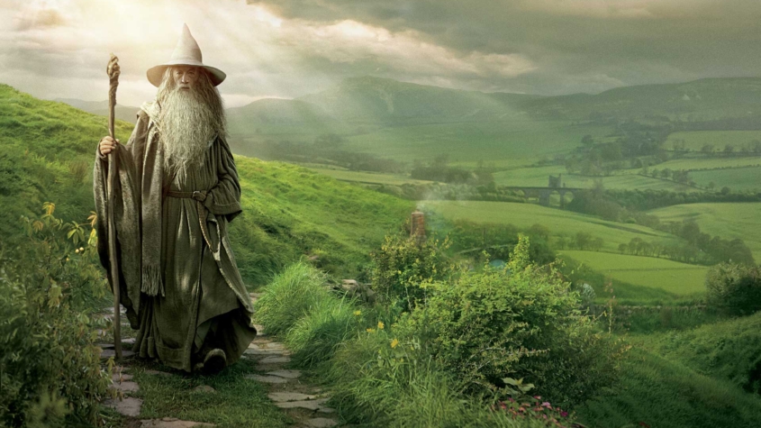 Gandalf the Grey from The Lord of the Rings stands on a stone path in the lush green hills of the Shire. Wearing his iconic pointed wizard hat and gray robes, he holds his wooden staff while overlooking the idyllic countryside. Sunlight breaks through clouds in the background, illuminating the pastoral landscape of rolling hills and distant fields that stretches toward the horizon. The scene captures Gandalf as the wise mentor figure at the beginning of the hero's journey.