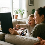 013- TMB Blog Post- Featured-Image-2 Parent and young child sitting together on a couch, both smiling and engaged while watching educational content on a tablet in a warm, sunlit living room