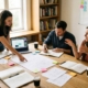 Three educators collaborating around a table covered with notebooks, sketches, and sticky notes in a bright office with bookshelves and a whiteboard, actively discussing and planning together