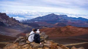 Two people sitting on volcanic rocks overlooking Haleakalā crater in Maui, with dramatic red and brown cinder cones and clouds in the valley below
