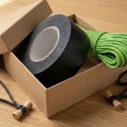Kraft paper gift box containing a roll of black gaff tape, coiled bright green 550 paracord, and bamboo Bongo Ties with elastic loops on a wooden surface