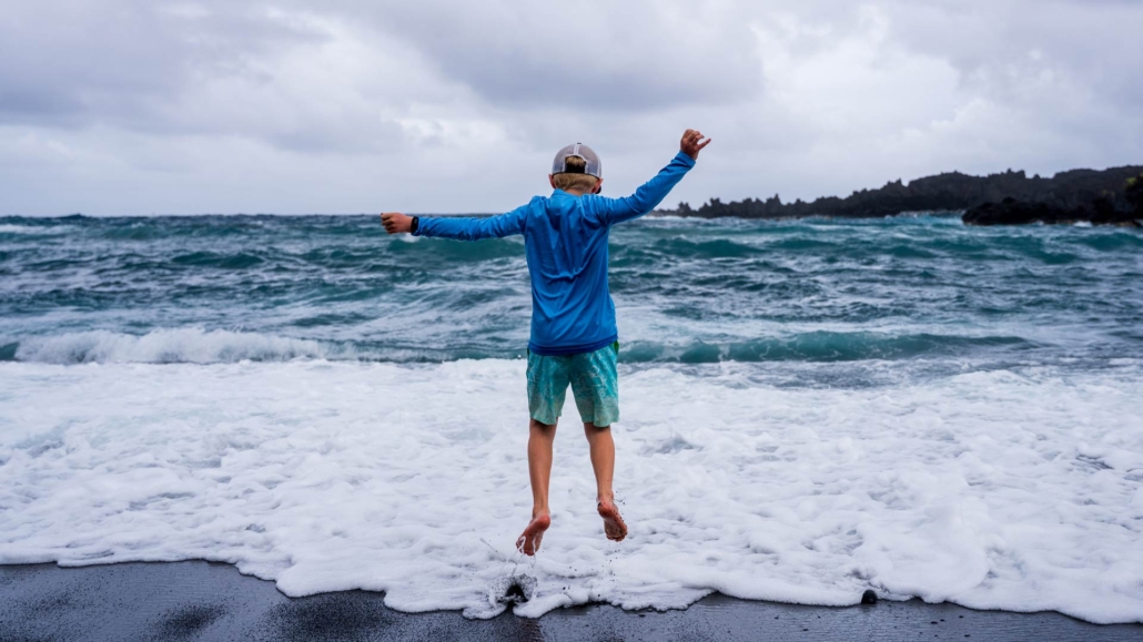 Child jumping joyfully on a black sand beach in Hawaii, with turquoise ocean waves and volcanic coastline in the background